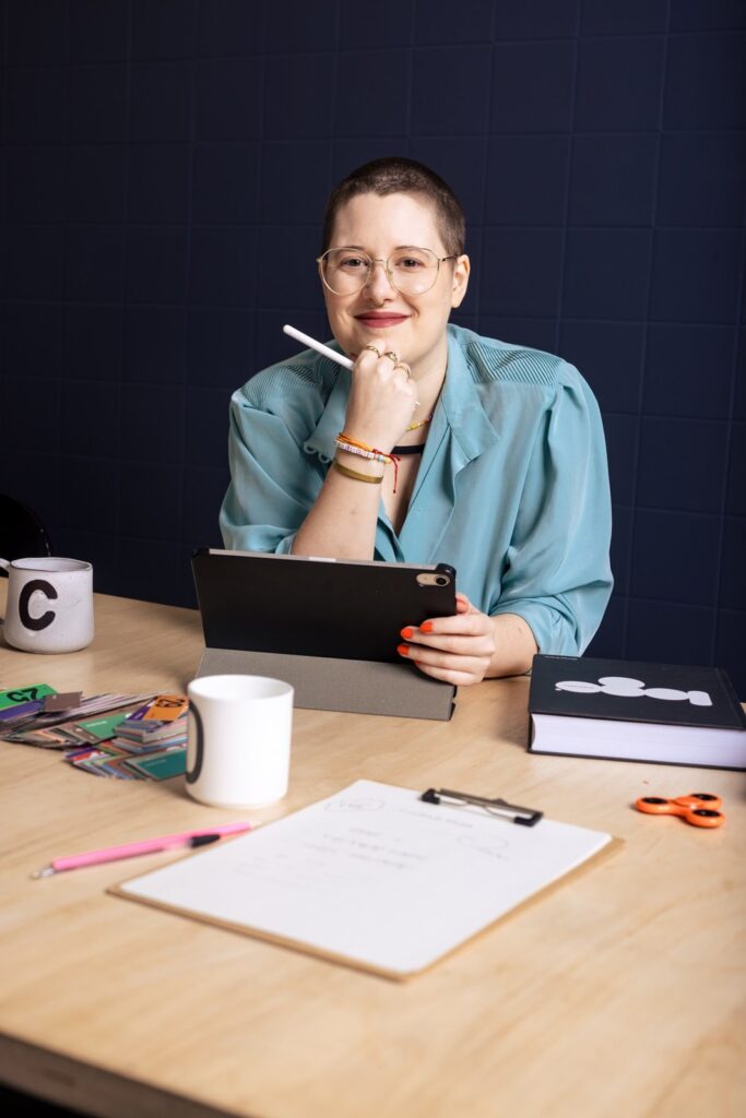 Porträt einer lächelnden Frau mit kurzem Haar und Brille, die an einem Schreibtisch mit iPad, Farbmustern und Notizen sitzt – symbolisiert kreative Markenarbeit.